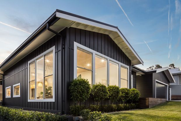 Exterior view of a contemporary single-storey home with dark vertical cladding, a gabled roof, contrasting light eaves, large white-framed windows, and neatly landscaped hedges. A connected garage or carport structure is visible to the side, with neighboring homes in the background.