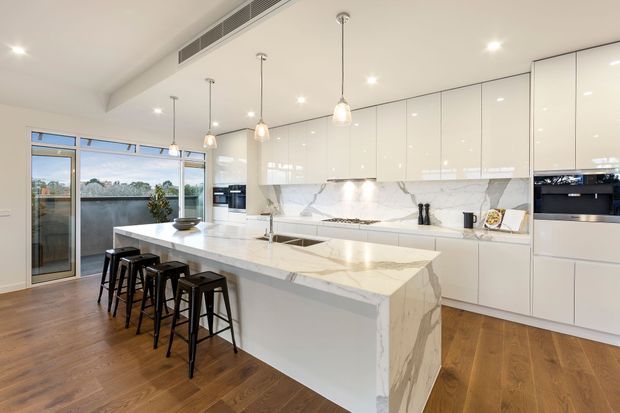 Bright contemporary kitchen with a large marble-look island and four black bar stools, glossy white full-height cabinetry, integrated appliances, pendant lighting, timber flooring, and a glass door opening to a balcony or terrace with a water view.