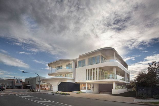 Street-level exterior view of a large contemporary multi-storey building with a sculptural curved roofline, rounded balconies, extensive glazing, white rendered facade, vertical window fins, and integrated ground-level garages. The building sits prominently on an urban street corner with neighboring commercial or residential structures visible nearby.