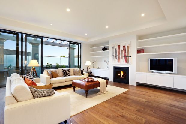 Bright, spacious contemporary living room with two cream sofas, a leather ottoman coffee table, light rug, timber flooring, built-in white shelving and media cabinetry, and a modern fireplace. Full-height black-framed glazing opens to an outdoor area, while recessed ceiling lights provide even illumination. Abstract artwork and restrained furnishings create a refined, inviting atmosphere.