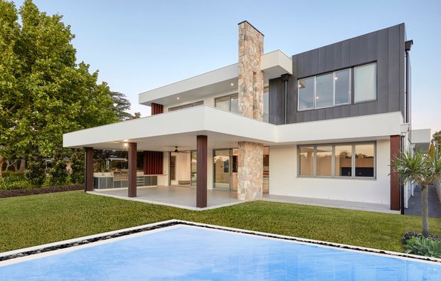 Exterior view of a contemporary two-storey residence with strong horizontal roof and balcony lines, white rendered walls, dark vertical metal cladding, extensive glazing, and a prominent natural stone chimney. A covered outdoor entertaining area and swimming pool sit in the landscaped front garden.