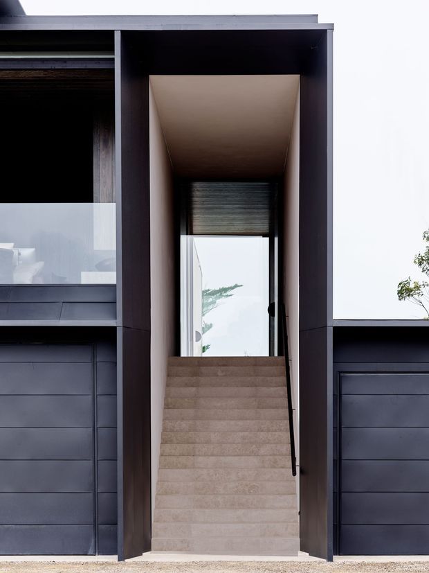 Front-facing exterior view of a contemporary multi-level home entrance. A tall recessed entry is framed by dark charcoal cladding, with a wide flight of pale stone or concrete stairs leading to a glazed door. Large windows and integrated dark garage doors flank the entry, creating a strong vertical architectural composition.