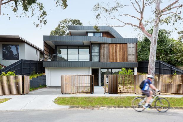Street-facing view of a contemporary multi-level residence with dark metal cladding, a prominent warm timber-screened upper facade, extensive glazing, and a compact front garden enclosed by timber and dark fencing. The house has a strong horizontal composition with a pitched roof element and a recessed ground-floor entry and garage-like opening. A cyclist passes in the foreground.