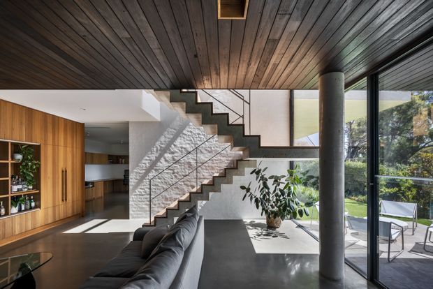 A contemporary open-plan living space with a dramatic floating zigzag staircase, dark timber-lined ceiling, polished concrete flooring, and extensive floor-to-ceiling glazing opening to a garden terrace. Warm timber built-in cabinetry and shelving define the kitchen and living area, while a substantial concrete column adds a strong structural element. The room is bright and spacious with a refined natural-material palette.