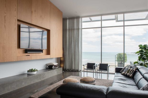Contemporary living room with a large built-in timber entertainment wall and grey concrete-like console, a black sectional sofa, and full-height glazed doors opening to a beachfront view. Sheer curtains, a textured rug, and restrained furnishings create a refined, airy coastal atmosphere.