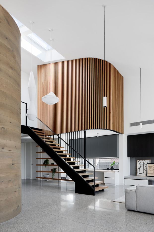 Bright, double-height contemporary interior featuring a sculptural open-riser staircase with black steel stringers and balustrade, warm timber treads, and a large curved vertical timber-slat feature wall. A rounded timber-clad structural volume frames the left side, while white walls, polished concrete or terrazzo flooring, minimalist pendant lights, and streamlined dark-and-white cabinetry create a clean, refined open-plan living area. A skylight and high-level glazing bring in abundant natural light.