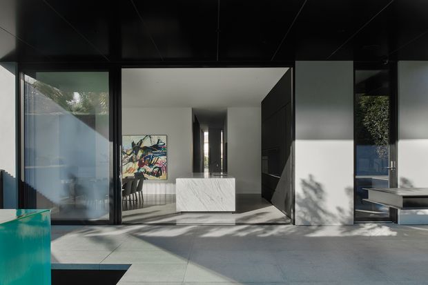 A contemporary open-plan interior viewed through large black-framed glazing, with a minimalist white kitchen island featuring a marble-look surface, dark full-height cabinetry, polished concrete or large-format grey flooring, and a sculptural colorful artwork in the adjoining living area. Strong indoor-outdoor connection, sharp geometric lines, and dramatic sunlight and shadows define the scene.