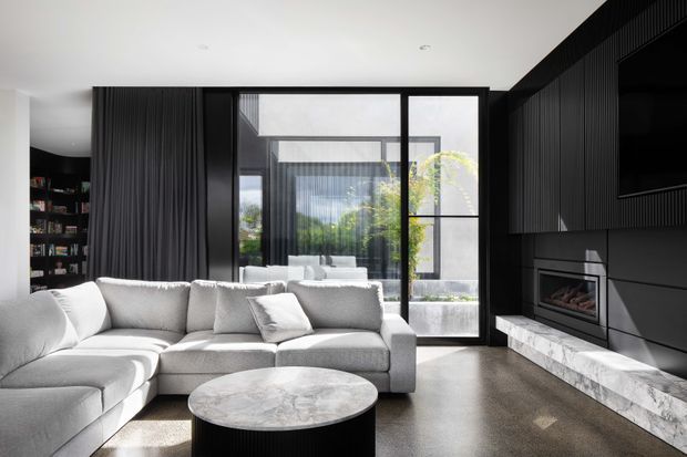 Contemporary living room with a large light-grey sectional sofa arranged around a round marble coffee table. The room features dark vertically ribbed built-in cabinetry and wall panelling, a recessed fireplace with a marble hearth, black-framed floor-to-ceiling glazing, and a leafy courtyard visible beyond. The palette is monochromatic with strong black-and-white contrast and abundant natural light.