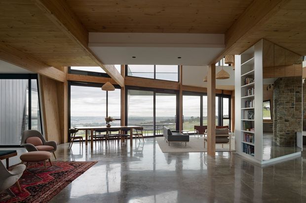 Open-plan contemporary living and dining space with exposed timber beams and a timber-lined ceiling, polished concrete flooring, extensive floor-to-ceiling glazing, and a built-in shelving and fireplace wall. Mid-century-inspired furniture, pendant lights, and a patterned rug furnish the room, with a broad landscape view beyond the windows.