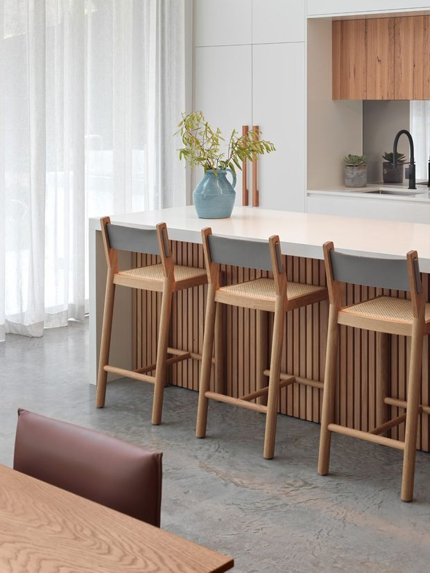 Bright, contemporary kitchen and dining area featuring a long white island with integrated seating, a vertical timber-slat island front, timber cabinetry, and a dark faucet and sink. Four timber bar stools with woven seats line the island. The space has a polished concrete floor, sheer white curtains, pale cabinetry, and a dining table with a leather chair in the foreground. A blue ceramic vase and indoor plants add restrained colour.