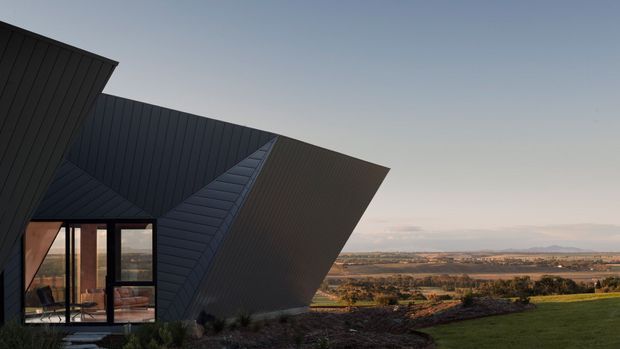 Exterior architectural photograph of a sharply angular, sculptural dark-clad house set on an elevated rural site. The faceted roof and wall planes create a dramatic geometric form, with floor-to-ceiling glazed doors opening onto a small terrace and expansive agricultural landscape beyond.