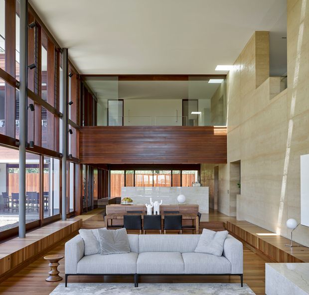 A spacious double-height contemporary living and dining area with floor-to-ceiling glazing along one side, warm timber framing and wall panelling, a large pale sectional sofa, dining table, and a mezzanine or upper-level walkway overlooking the room. A substantial pale stone or concrete feature wall and built-in hearth-like volume contrast with the timber. The space is bright, refined, and strongly connected to the exterior through expansive windows.