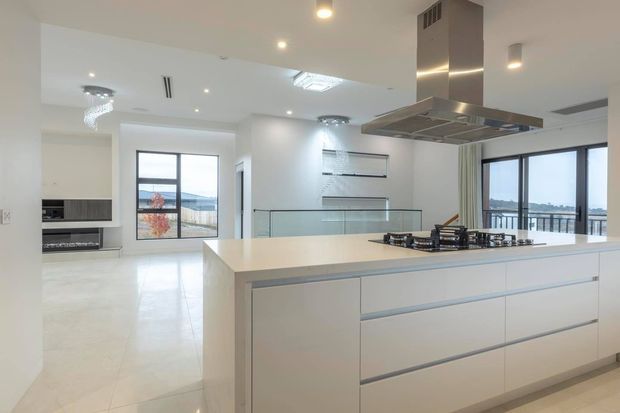 Bright, open-plan contemporary kitchen with a large white island, integrated cabinetry, gas cooktop and oversized stainless-steel rangehood. The space has glossy light flooring, white walls, recessed lighting, glass balustrading and expansive windows overlooking a waterfront or city-edge view.