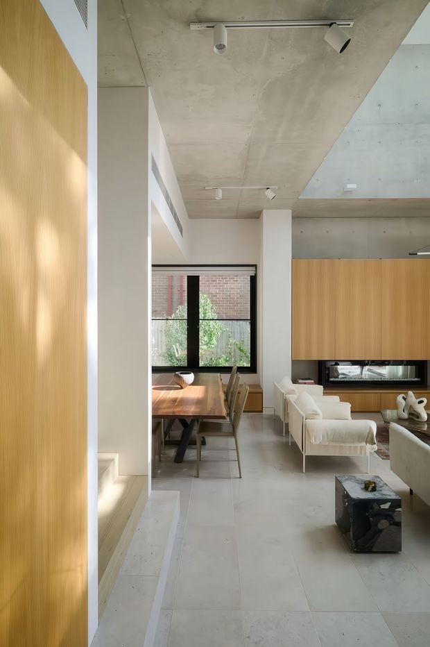 A refined open-plan living and dining interior with a minimalist, contemporary aesthetic. The space features a polished concrete ceiling with exposed services and track lighting, pale stone or concrete-look flooring, white walls, warm vertical timber wall panelling, and a large black-framed window. A timber dining table sits beside a light upholstered lounge area, with a long built-in timber media wall and fireplace forming the focal point.
