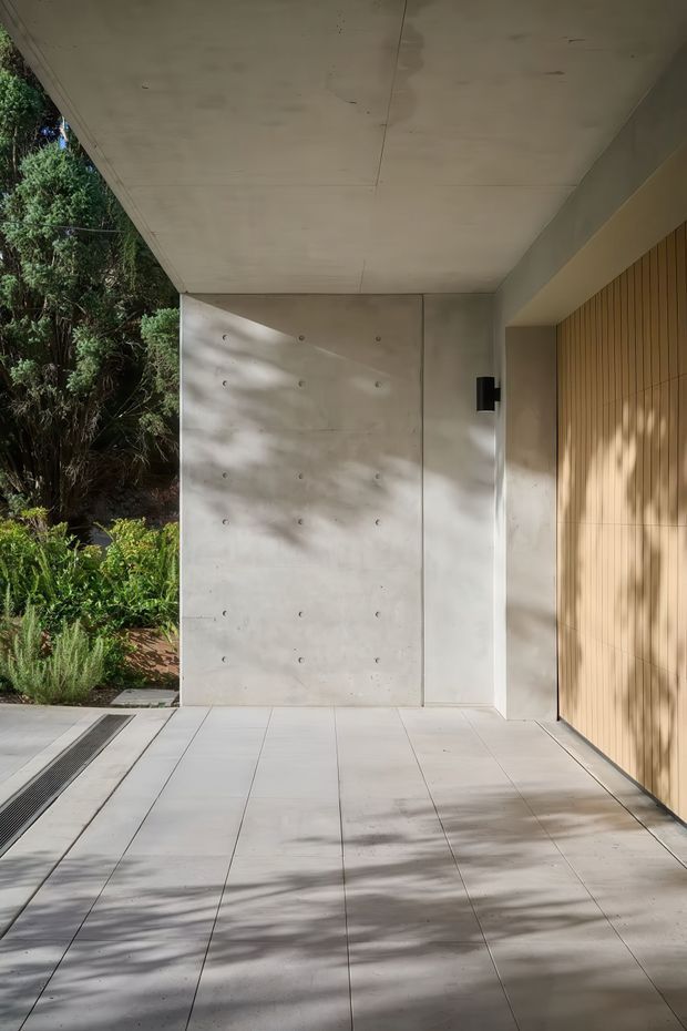 A minimalist covered exterior passage or entry area with large pale concrete wall and ceiling panels, a warm vertical timber-clad door or garage door, and large light rectangular floor tiles. Dense landscaped greenery borders the open side, with strong dappled sunlight and shadows across the surfaces.