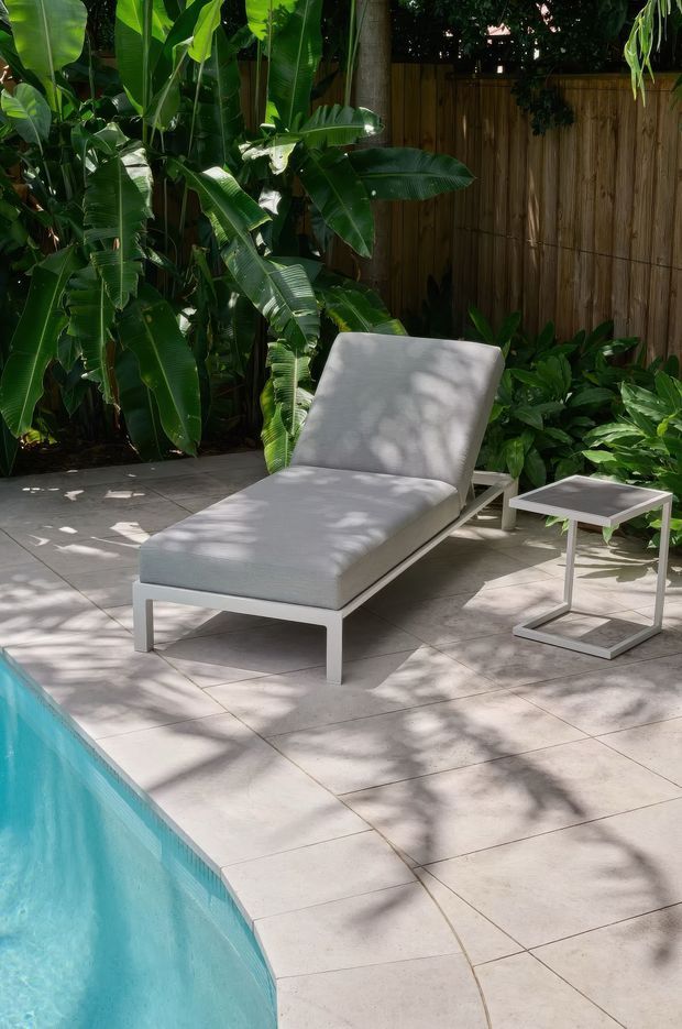 A sunlit tropical poolside patio with a white outdoor chaise lounge and matching side table on large pale tiled paving. Dense banana and broad-leaf planting borders a warm timber privacy fence, while the turquoise swimming pool occupies the foreground.