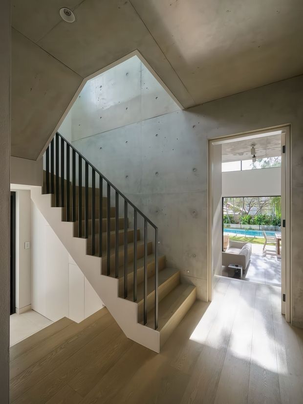 Interior view of a contemporary residential stairway and landing. A straight-flight staircase with pale timber treads and dark slender vertical balusters rises beside a raw exposed concrete feature wall. Pale timber flooring continues through the landing, with white built-in storage beneath the stairs. A doorway opens toward a bright outdoor lounge and swimming pool beyond.