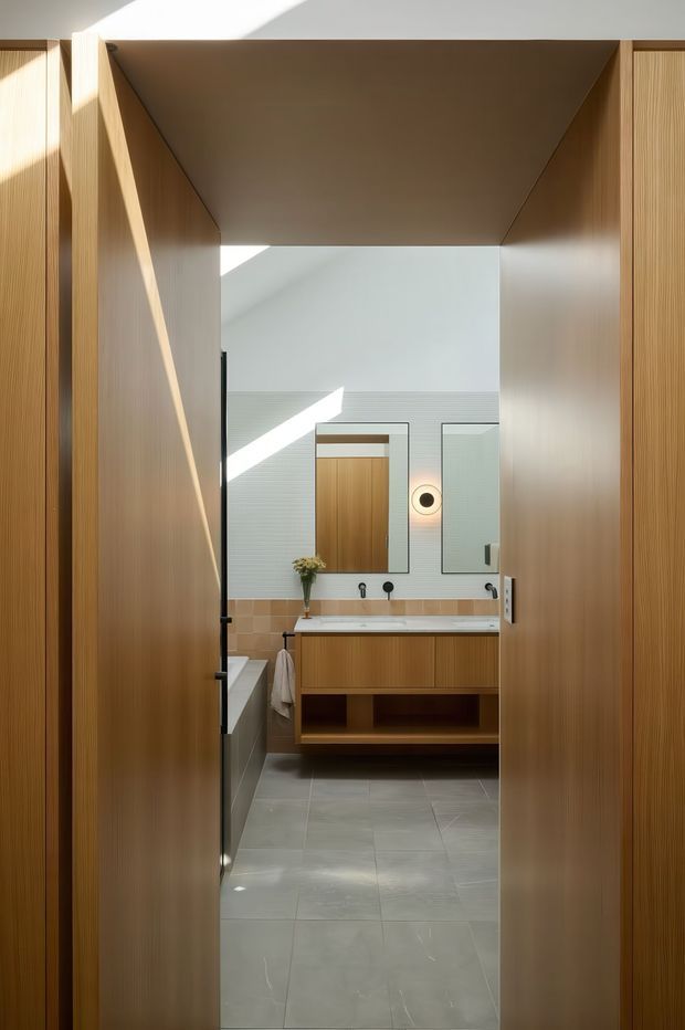 A contemporary bathroom viewed through a partially open timber doorway. The room features a floating timber vanity with integrated basin, wall-mounted black tapware, a large mirror, grey floor tiles, pale green-grey wall tiles, and a warm tan tiled backsplash. Strong natural light creates geometric shadows across the timber joinery and walls.