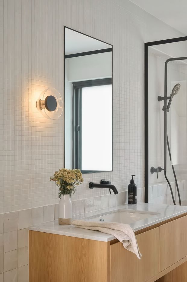 A refined contemporary bathroom vanity with a wall-mounted black tap, integrated white basin, light timber cabinetry, white tiled walls, and a large rectangular mirror. A black-framed glass shower enclosure with matching dark fixtures is visible at right. A round wall sconce provides warm illumination, while a vase and folded towel add subtle styling.
