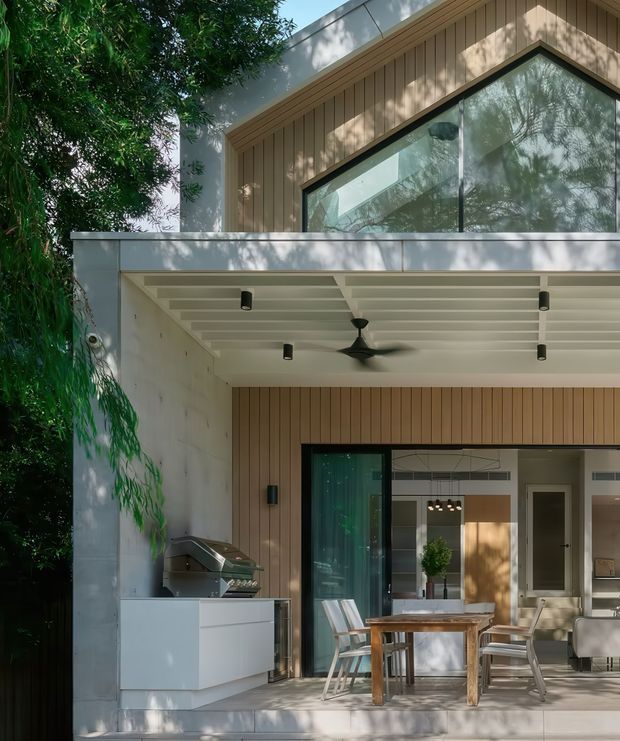 Modern two-storey residential exterior featuring a covered outdoor entertaining area with a built-in barbecue, outdoor dining table and chairs, ceiling fan, recessed lighting, and large glazed doors opening into the interior. The upper facade combines vertical timber cladding with a prominent angular gable window, while white rendered or masonry elements frame the structure. Lush vegetation partially surrounds the home.