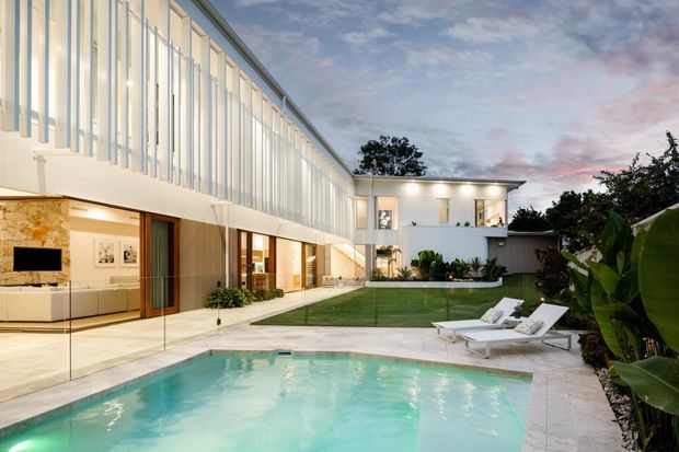 Contemporary two-storey residence arranged around a landscaped courtyard and swimming pool. The home features extensive floor-to-ceiling glazing, a long upper-level facade with vertical white fins, warm timber cladding, pale stone surfaces, and a flat roof. The pool terrace includes white sun loungers and lush tropical planting, creating a refined indoor-outdoor setting at dusk.
