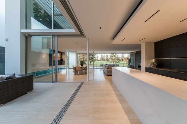 A bright, expansive contemporary open-plan interior viewed from the entry, with a large white kitchen island in the foreground, dark full-height cabinetry and integrated media joinery along the right wall, and a dining and living area extending toward full-height glazed doors overlooking a landscaped yard. Pale stone-look flooring, minimalist recessed linear lighting, extensive glazing, and clean architectural lines create a refined indoor-outdoor connection.