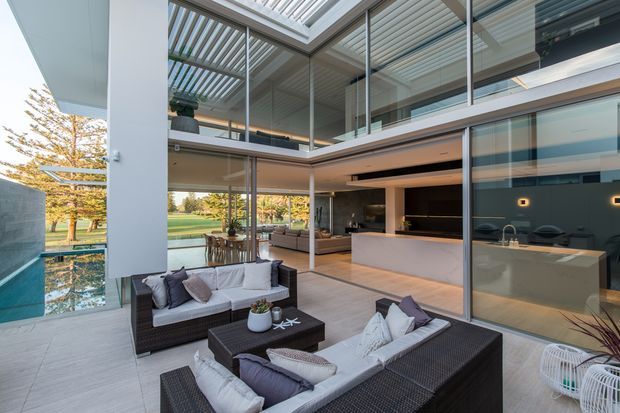 A sophisticated contemporary home interior opening onto a covered outdoor terrace, with extensive floor-to-ceiling glazing, a double-height living area, outdoor lounge furniture, and a sleek kitchen with a large pale island. A reflecting pool and landscaped garden are visible beyond the glass. The space uses restrained neutral tones, strong horizontal lines, and a louvred roof structure that brings in filtered daylight.