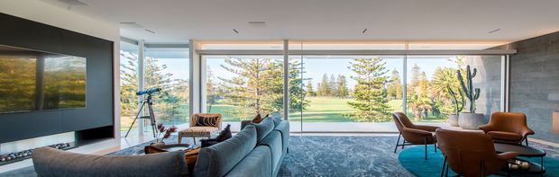 A spacious contemporary living room with a large sectional sofa, lounge chairs, coffee tables, and a wall-mounted television. Floor-to-ceiling glazing opens views to a landscaped garden, while a linear recessed ceiling light and dark feature wall add a refined modern character.
