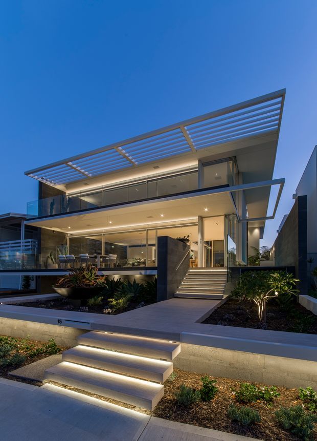 Twilight exterior photograph of a contemporary multi-level residence with extensive floor-to-ceiling glazing, projecting balconies, clean horizontal lines, and a large slatted roof canopy. Illuminated entry stairs lead through landscaped garden beds to the glass-fronted home, with warm interior and architectural lighting creating a refined modern appearance.