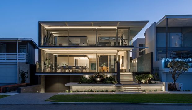 A contemporary multi-level residential house photographed at dusk, featuring strong horizontal lines, extensive floor-to-ceiling glazing, cantilevered upper levels, a flat roof, open balconies, and a recessed garage. Warm interior lighting reveals living spaces and planted terraces, while broad steps connect the landscaped front yard to the entry.