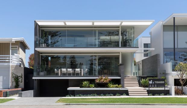 Contemporary two-storey residential façade with extensive floor-to-ceiling glazing, broad cantilevered flat roof slabs, glass balustrades, dark lower-level cladding, and a stepped entry staircase. The front garden includes built-in raised planters and restrained landscaping, with neighbouring modern homes visible on either side.