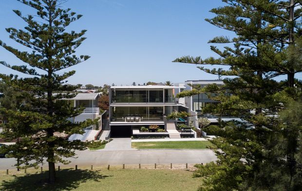 Exterior view of a contemporary multi-level residence framed by large mature conifer trees. The home features a rectilinear form, extensive floor-to-ceiling glazing, projecting balconies and terraces, light-coloured rendered or concrete surfaces, and a landscaped front approach.