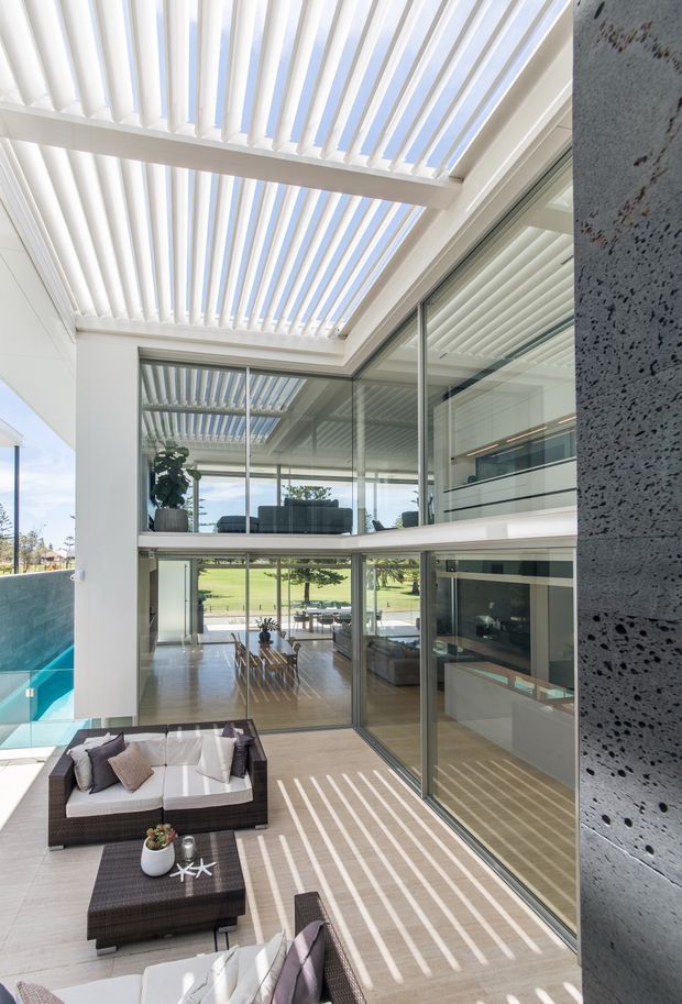 A bright, contemporary multi-level living space centered on a glazed internal courtyard or atrium. White louvred pergola-style roof panels cast strong linear shadows across pale tiled flooring and outdoor-style wicker seating. Full-height glass walls reveal an upper-level lounge, a lower dining and living area, and expansive views to a green landscape. A dark textured concrete or stone feature wall frames the right side, while a turquoise pool is visible at the left edge.