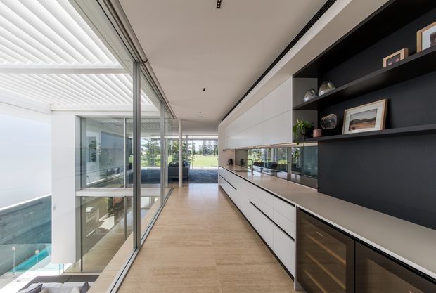 A bright, long contemporary interior corridor or open-plan living area with full-height glazing along the left side, overlooking a lower-level pool and outdoor space. The right side features extensive built-in white cabinetry, a reflective splashback, dark open shelving, and integrated glass-front storage. Pale timber flooring, concealed linear lighting, and clean rectilinear detailing create a refined minimalist interior.