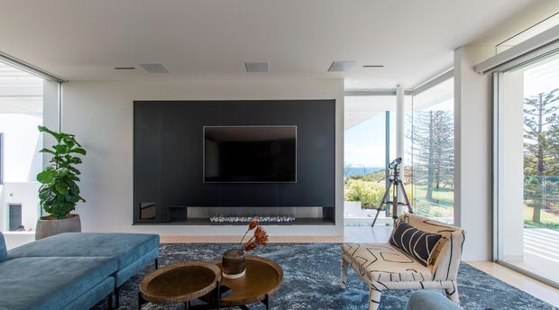Bright contemporary living room with expansive floor-to-ceiling glazing, a deep blue sectional sofa, patterned armchair, round nesting coffee tables, and a large television integrated into a dark feature wall with a long recessed media console. The room opens to a scenic coastal or landscaped outlook and is finished with a textured blue-grey rug and restrained modern furnishings.