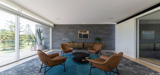 A refined contemporary living room with a low-profile seating arrangement centred on a round black coffee table. Brown leather lounge chairs sit on a deep teal rug, set against a dark grey textured feature wall with a slim console and artwork. Floor-to-ceiling glazing opens the room to a landscaped exterior, while a large internal glass opening is visible on the right. The space has a clean, minimalist composition with strong natural light and warm timber accents.