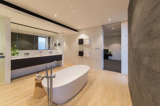 A spacious contemporary bathroom with a freestanding white bathtub, wall-mounted bath tap, floating dark vanity, large mirror, double basins, towel rails, recessed ceiling lights, and pale timber-look flooring. A textured grey stone feature wall frames the right side, while an open doorway leads to an adjacent darker room.
