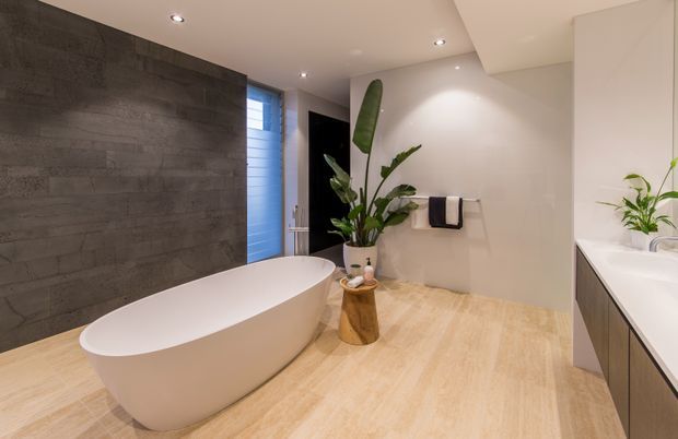 A spacious contemporary bathroom featuring a freestanding white oval bathtub, large-format dark grey wall tiles, pale wood-look floor tiles, a floating timber-toned vanity with white countertop, recessed ceiling spotlights, and indoor tropical plants. A blue-toned glazed door or window adds colour at the rear.