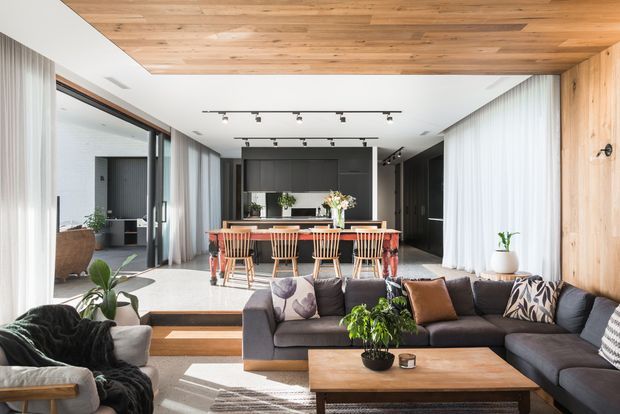 A bright open-plan contemporary living and dining space with a kitchen beyond. The room features a large charcoal sectional sofa, timber coffee table, timber dining table with spindle-back chairs, extensive floor-to-ceiling glazing with sheer curtains, and warm timber lining across the ceiling and one wall. Track lighting and dark kitchen cabinetry provide contrast, while indoor plants and neutral furnishings add a relaxed, refined atmosphere.
