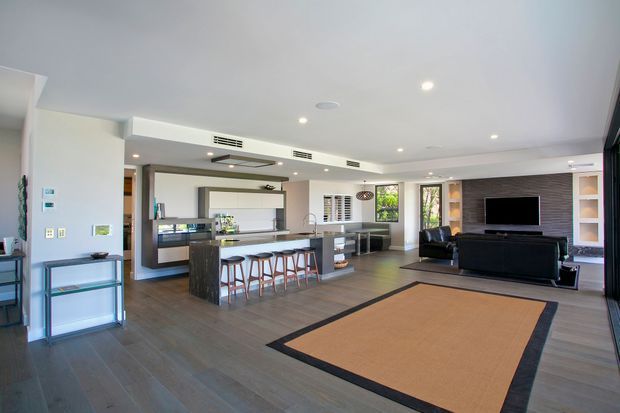 Open-plan contemporary living space combining a kitchen with a central island and bar seating, dining/work area, and a lounge with a wall-mounted television. The room has dark timber-look flooring, white walls and ceiling, recessed lighting, extensive glazing, dark built-in cabinetry, and a large neutral rug with a dark border.