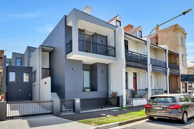 Street-facing view of a contemporary multi-storey townhouse inserted beside attached traditional terrace homes. The new dwelling has a dark grey and white rendered facade, projecting balconies with black metal balustrades, a small front garden and gated entry, while neighbouring homes feature rendered masonry, ornate detailing and pitched dormer roofs.