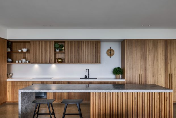 A contemporary kitchen viewed straight on, featuring extensive vertical timber battens and wood cabinetry, open display shelving, a large grey stone island with integrated seating, pale walls, recessed ceiling lights, and understated decorative objects and plants.