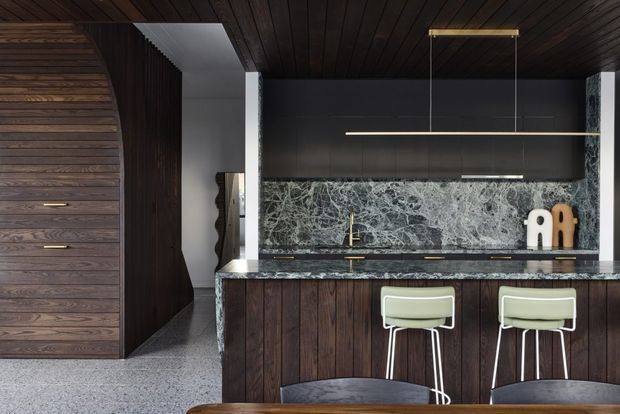 A sophisticated dark-toned kitchen with a peninsula or island clad in vertically grooved dark timber, a dramatic veined stone splashback and benchtop, integrated dark cabinetry, and two pale green upholstered bar stools. The room features a dark timber-lined ceiling, slim linear pendant lighting, polished metal tapware, and terrazzo-like flooring. A large timber-clad curved or paneled wall is visible to the left, with a passage opening beyond the kitchen.