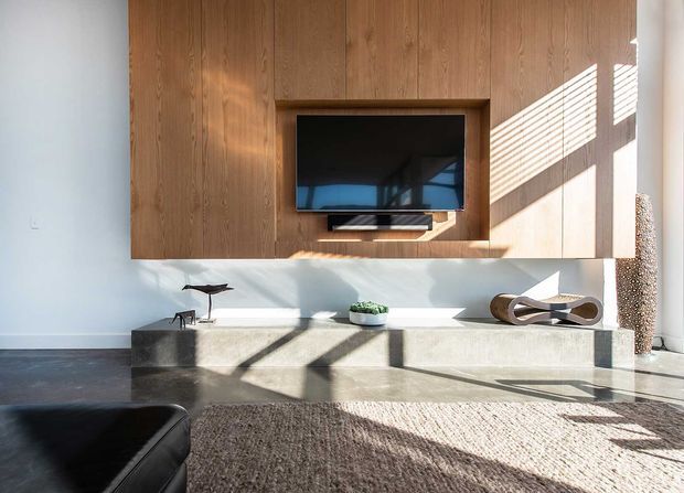 A bright, minimalist living room featuring a full-height warm timber media wall with an inset television, a long pale stone or concrete built-in console, and a low contemporary seating arrangement. Strong sunlight enters through horizontal blinds, casting graphic shadows across the wall, floor, and textured neutral rug. Sculptural decor and restrained furnishings create a calm, refined atmosphere.