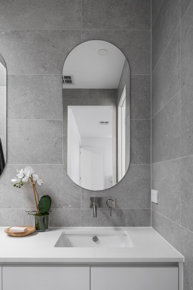 A compact contemporary bathroom vanity with large-format grey stone-look wall tiles, a white integrated basin and benchtop, wall-mounted chrome tapware, and an arched-edge oval mirror. White cabinetry sits below the vanity, while a small orchid adds a natural accent.