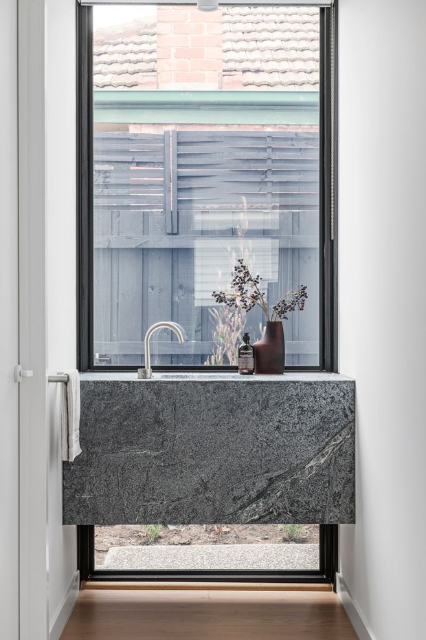 A compact contemporary bathroom vanity positioned in front of a large black-framed window. The floating vanity has a dramatic dark grey natural stone slab with prominent veining, paired with a simple curved chrome tap. White walls, timber flooring, and a muted grey outdoor fence visible through the window create a clean, refined composition.