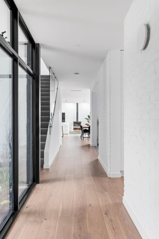 A bright, narrow residential hallway with light timber flooring, white-painted brick walls, and black-framed floor-to-ceiling glazing along the left side. A carpeted stairway rises near the entrance, while the corridor leads toward a dining and living area with contemporary furnishings. The space has a clean, minimalist aesthetic with strong contrast between white surfaces, pale wood, and black window frames.