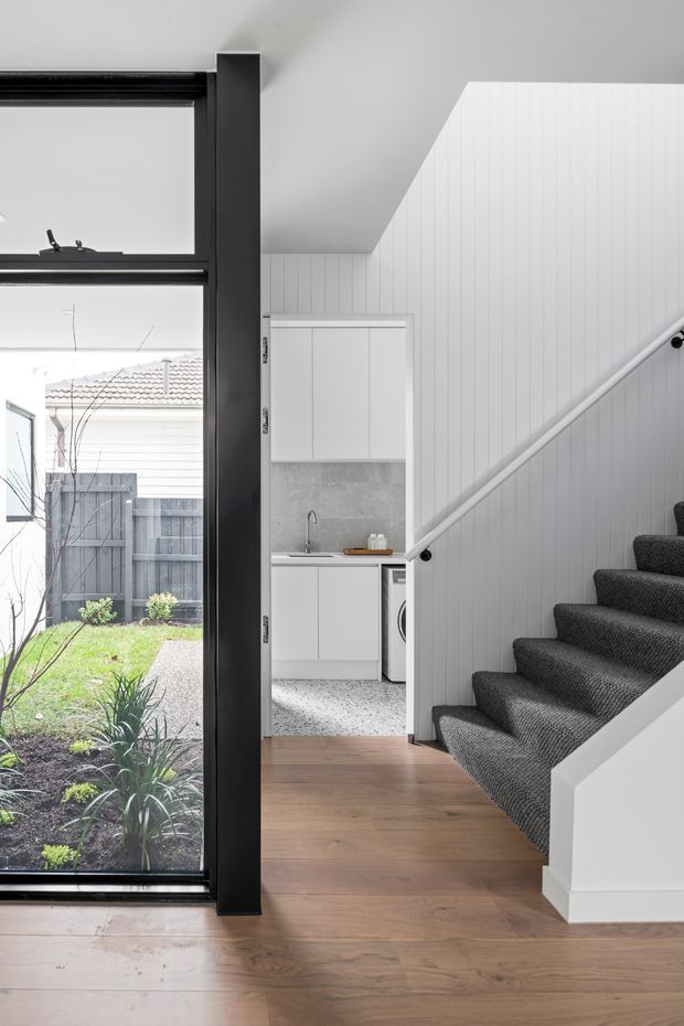 A bright contemporary residential interior showing a compact stairway beside a full-height black-framed window. The stairs have dark grey carpeted treads, a white painted balustrade and wall clad in vertical white panelling, with a simple white handrail. Beyond the stair area is a small white laundry or utility space with upper cabinets, a grey stone-look splashback, sink and washing machine. Warm timber flooring runs through the foreground, and a small planted courtyard is visible through the glazing.