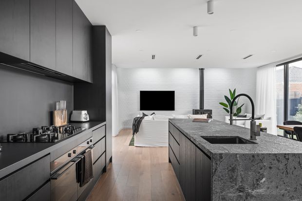 Open-plan contemporary kitchen and living area viewed from the kitchen. The kitchen features dark matte cabinetry, integrated appliances, a gas cooktop, and a large stone island with a sink and curved tap. Beyond it is a living area with a white sofa, wall-mounted television, white painted brick feature wall, fireplace flue, timber flooring, and expansive glazed doors or windows with blinds.