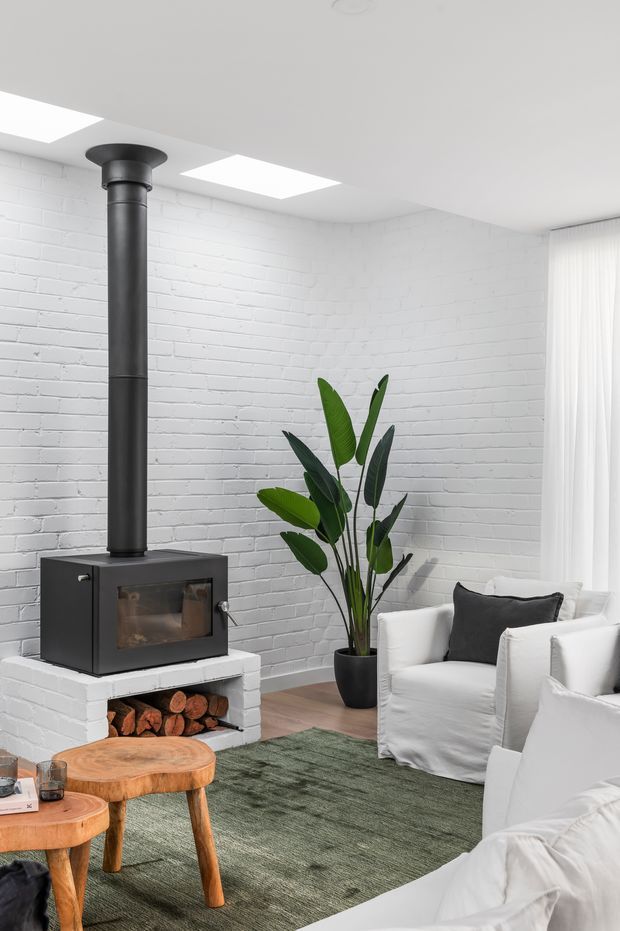Bright contemporary living room with white-painted brick walls and ceiling, a black freestanding wood-burning stove with exposed flue, white upholstered armchairs and sofa, a large leafy potted plant, green area rug, and rustic natural timber stump tables. Rectangular ceiling lights and sheer curtains contribute to a clean, airy interior.